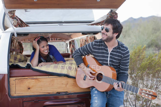 Hombre adulto medio tocando la guitarra en la parte trasera de la caravana, sonriendo - foto de stock