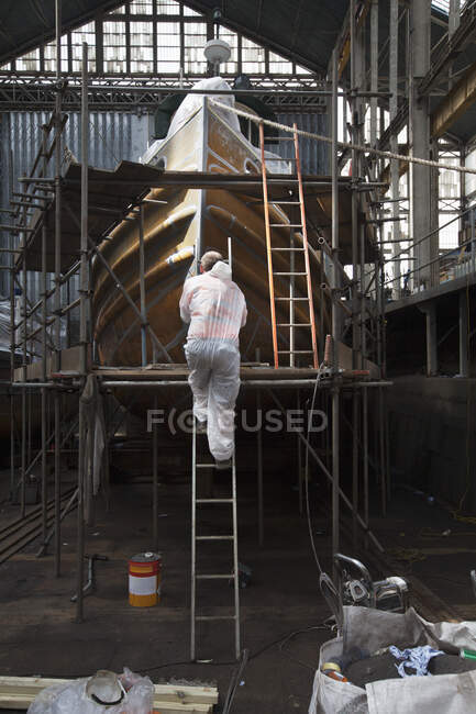 Worker climbing ladder to spraypaint boat in shipyard — Stock Photo