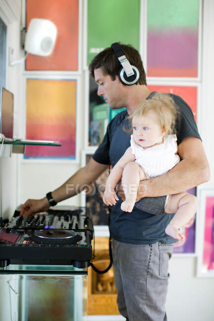 Hombre adulto medio cocinando, escuchando auriculares y llevando a su hija bebé - foto de stock