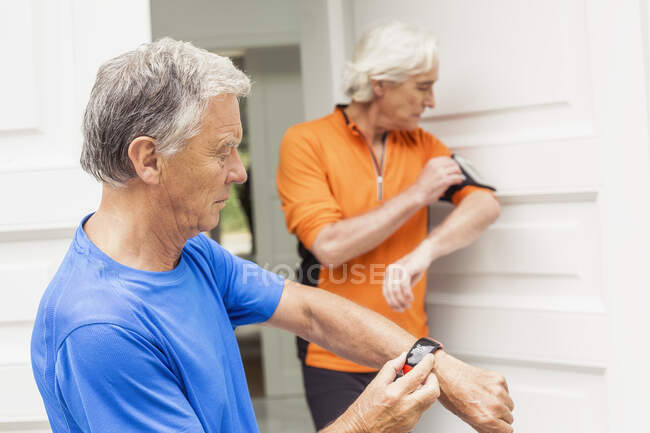 Two senior male runners at front door synchronising smartwatch and smartphone — Stock Photo