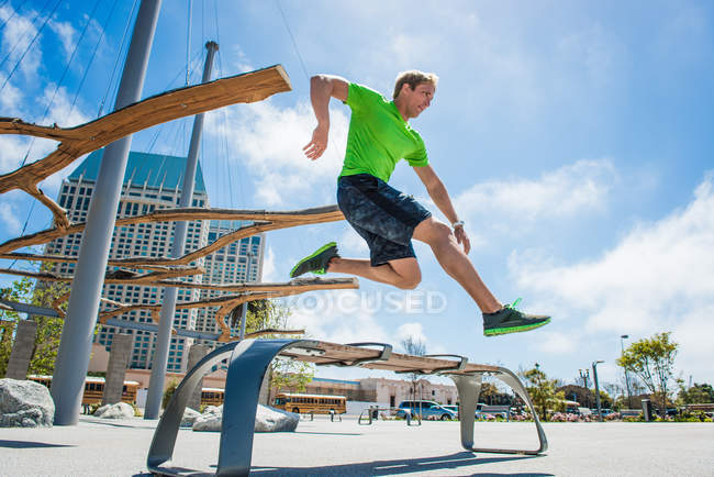 Joven saltando sobre el banco del parque en la ciudad - foto de stock