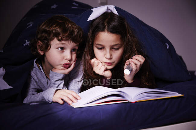 Irmão e irmã sob edredão leitura livro por tocha — Fotografia de Stock