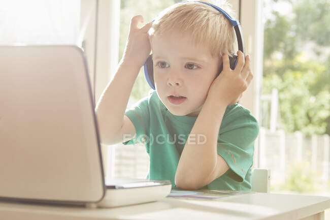 Niño en el escritorio usando el ordenador portátil y escuchando los auriculares - foto de stock