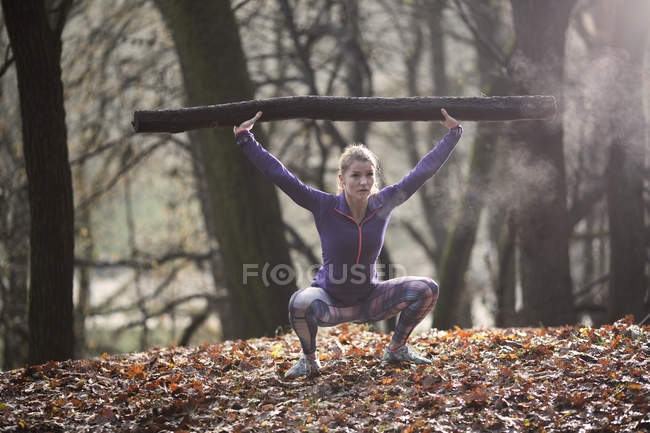 Front view of young woman squatting in forest arms raised holding tree branch — Stock Photo