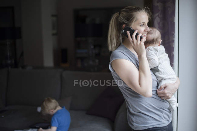 Mother holding baby boy, speaking on smartphone, while son uses digital tablet — Stock Photo