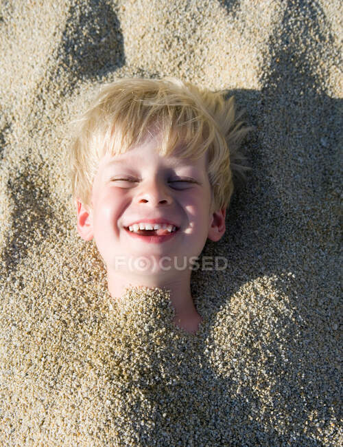 Niño enterrado en la arena riendo - foto de stock