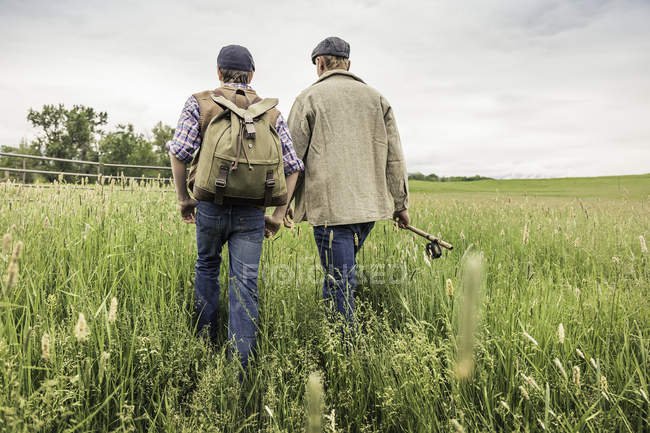 Visão traseira de homem e menino vestindo tampas planas carregando mochila e vara de pesca andando na grama alta — Fotografia de Stock