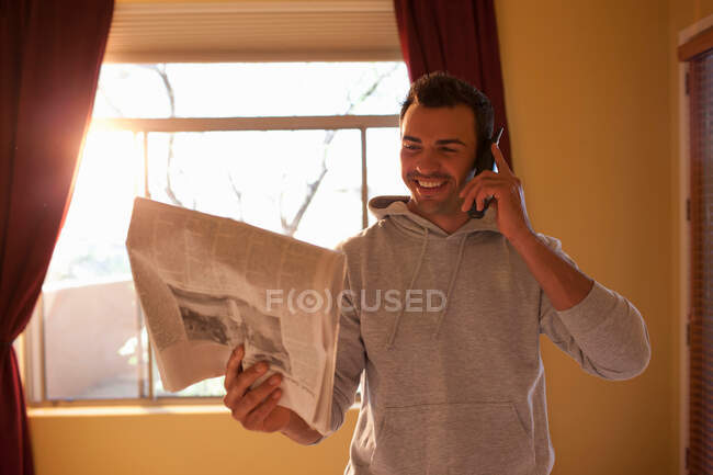 Молодий чоловік дивиться на газету і використовує мобільний телефон в готельному номері, посміхаючись — стокове фото