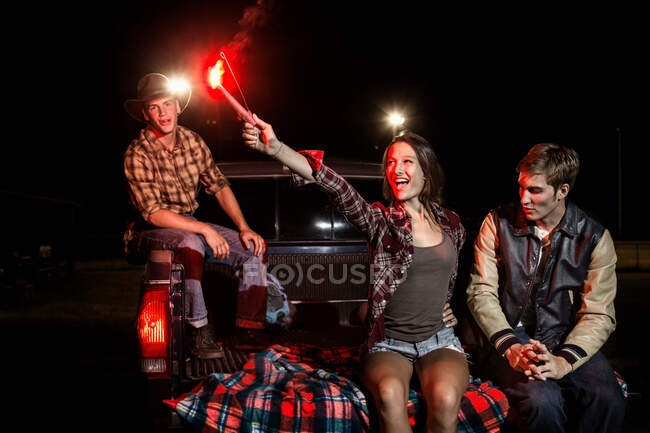 Three friends sitting on tailgate of car at night, girl holding sparkler — Stock Photo