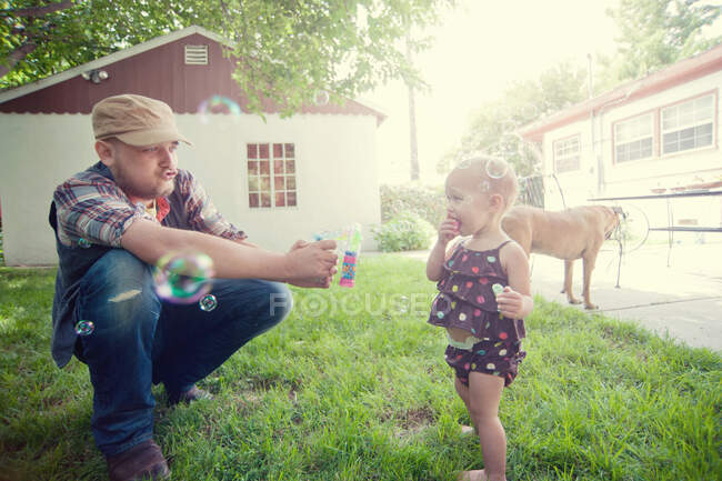 Батько і дитина дочка грають з бульбашками в саду — стокове фото