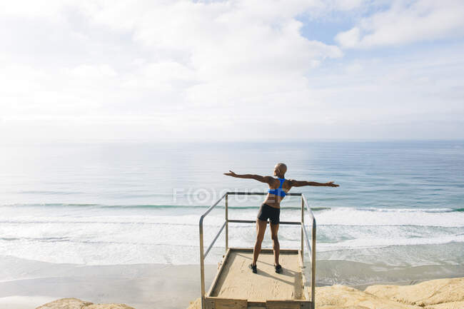 Young woman standing on look out point beside sea, arms raised, rear view — Stock Photo