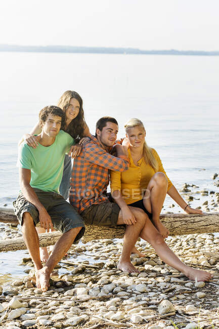 Friends sitting on driftwood next to shoreline looking at camera smiling, Schondorf, Ammersee, Bavaria, Germany — Stock Photo