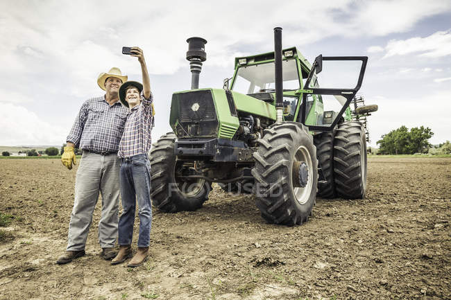 Фермер и внук делают селфи на смартфоне рядом с фермерским трактором — стоковое фото