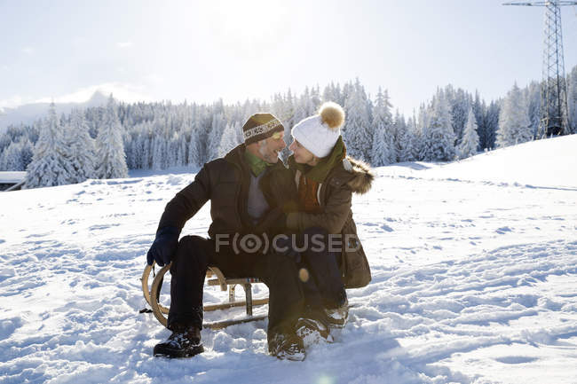 Senior couple on snowy landscape sitting on sledge face to face smiling, Sattelbergalm, Tyrol, Austria. - foto de stock