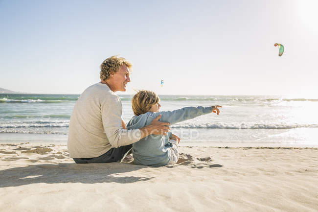 Father and son sitting on beach looking away pointing — Stock Photo