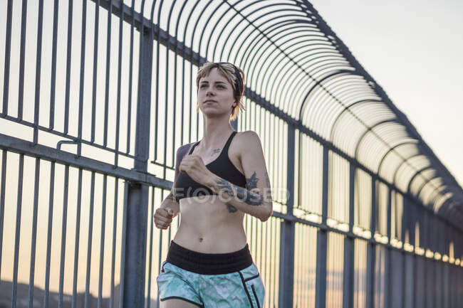 Молодая татуированная женщина бежит по мосту с закатом позади — стоковое фото