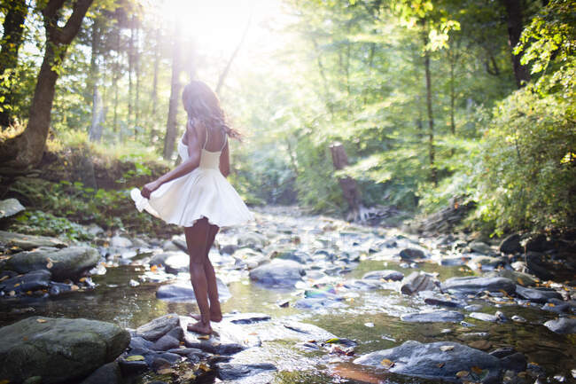 Glamorous young woman wearing white dress stepping over forest river rocks — Stock Photo