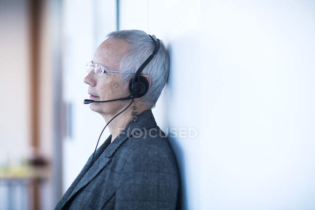 Vista lateral de una mujer madura apoyada en la pared con auriculares telefónicos mirando hacia otro lado - foto de stock