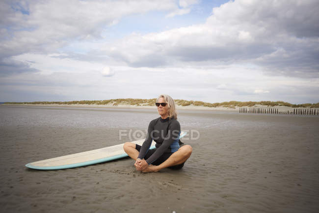 Senior mulher relaxante na areia, prancha de surf ao lado dela — Fotografia de Stock