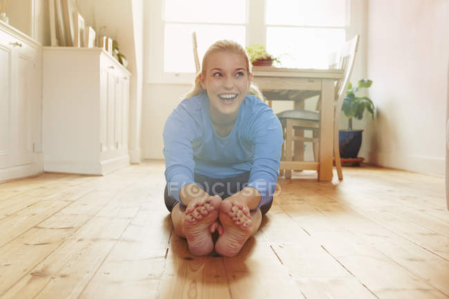 Молодая женщина, сидящая на полу, наклоняясь вперед, касаясь пальцев ног — стоковое фото
