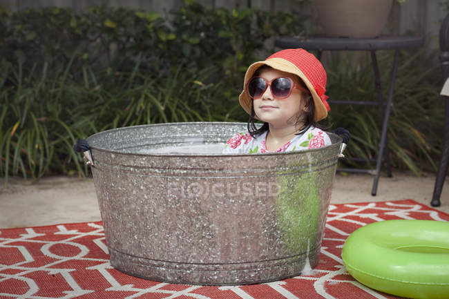 Retrato de niña con sombrero de sol y gafas de sol sentada en baño de burbujas en el jardín - foto de stock