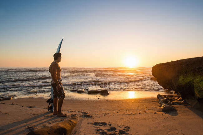 Человек, несущий доску для серфинга на скалистом пляже — стоковое фото