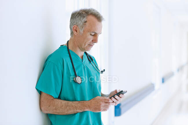 Cirujano de pie en el pasillo mirando el teléfono inteligente - foto de stock