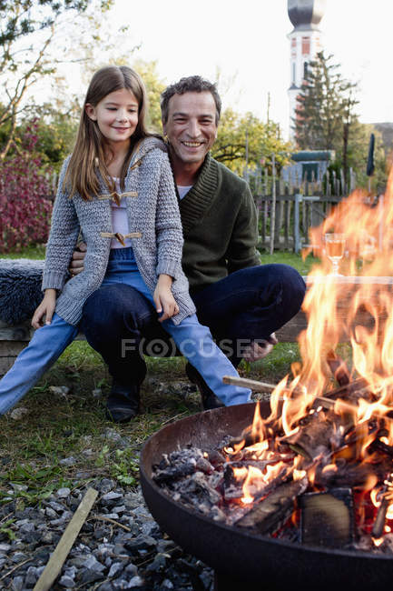 Девушка сидит на коленях отца в саду и смотрит на огненную яму — стоковое фото