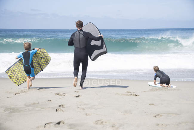 Rückansicht von Vater und zwei Söhnen, die mit Bodyboards am Strand, Lagunenstrand, Kalifornien, USA laufen — Stockfoto