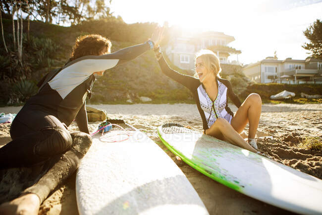 Coppia seduta sulla spiaggia, mani battenti in alto cinque, tavole da surf accanto a loro — Foto stock