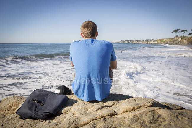 Молодий чоловік сидить біля скелі і дивиться на море ззаду. — стокове фото