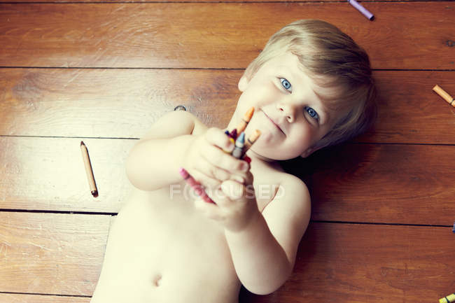 Vista de alto ângulo do menino deitado de costas no chão de madeira segurando lápis de cor, olhando para a câmera — Fotografia de Stock
