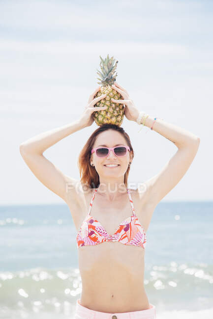 Retrato de mulher adulta média na praia, usando óculos de sol, segurando abacaxi na cabeça — Fotografia de Stock