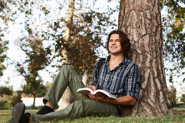 Молодий чоловік, сидячи проти дерева, тримає книгу, дивиться в очі посміхаючись — стокове фото