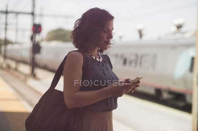 Mulher adulta média esperando na estação de trem, segurando smartphone — Fotografia de Stock