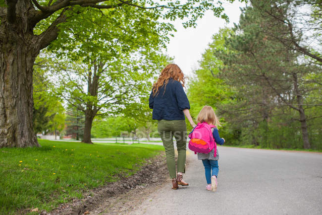 Rear view of mother and daughter holding hands walking down road through woods — Stock Photo