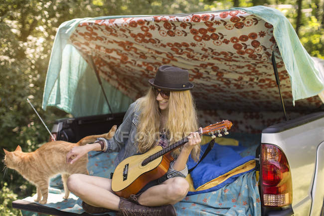 Young woman petting cat and playing ukulele whilst camping in pick up boot — Stock Photo