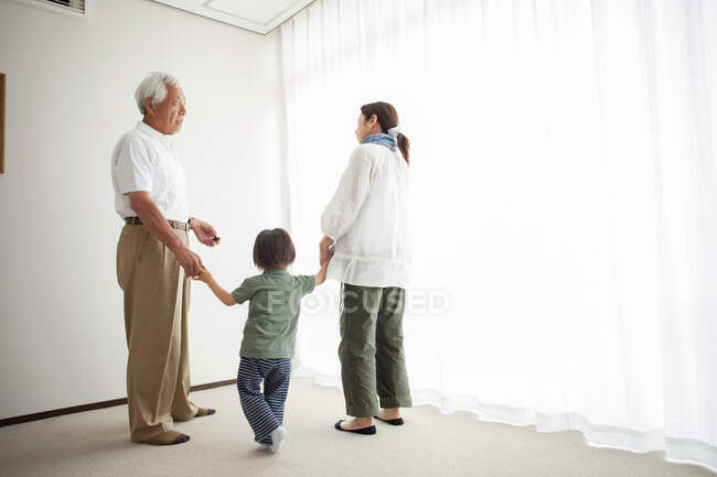 Сім'я трьох поколінь, що стоїть біля вікна, тримається за руки — стокове фото