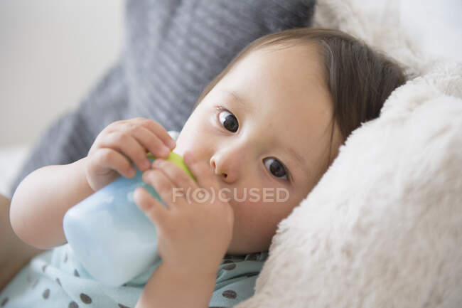 Retrato del bebé niño en el sofá bebiendo de la taza del bebé - foto de stock