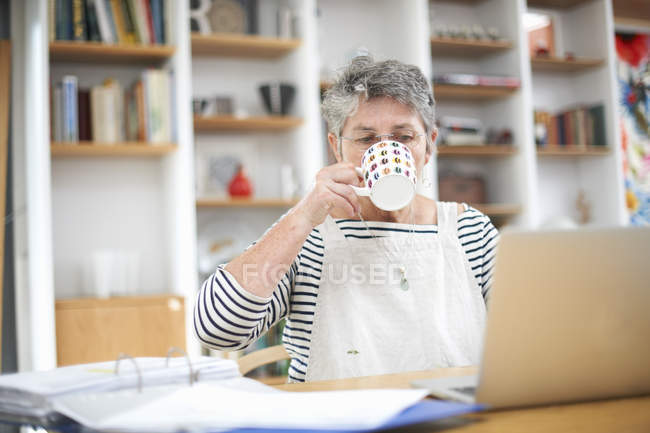 Mujer mayor en casa, usando laptop, bebiendo bebida caliente - foto de stock