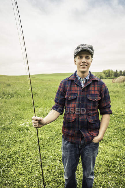Молодой человек в поле в чековой рубашке и плоской кепке держит удочку, глядя на камеру улыбаясь — стоковое фото