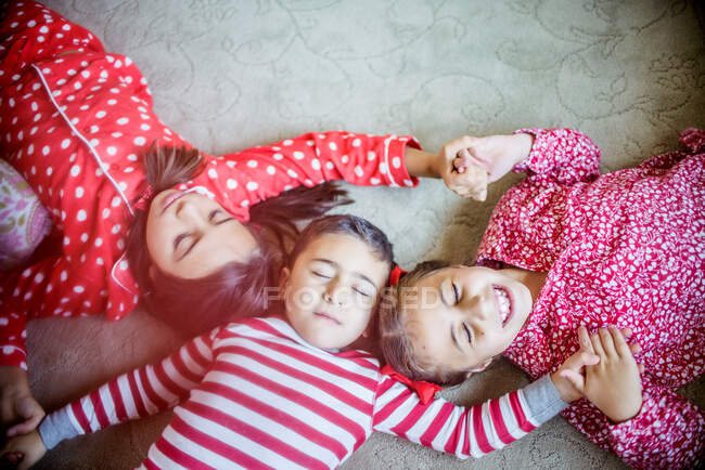 Overhead view of children wearing pyjamas lying on carpet holding hands eyes closed — Stock Photo
