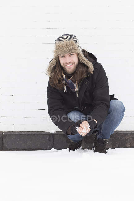 Jeune homme accroupi neige couvert rue faisant boule de neige — Photo de stock