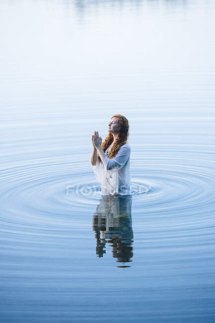 Young woman standing in lake ripples with eyes closed and hands together — Stock Photo