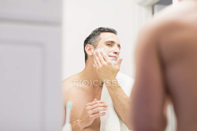 Середній дорослий чоловік, дивлячись у дзеркало, наносить піну для гоління обличчям, вид ззаду — стокове фото