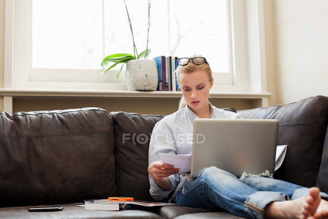 Jovem mulher sentada no sofá com laptop e papéis — Fotografia de Stock