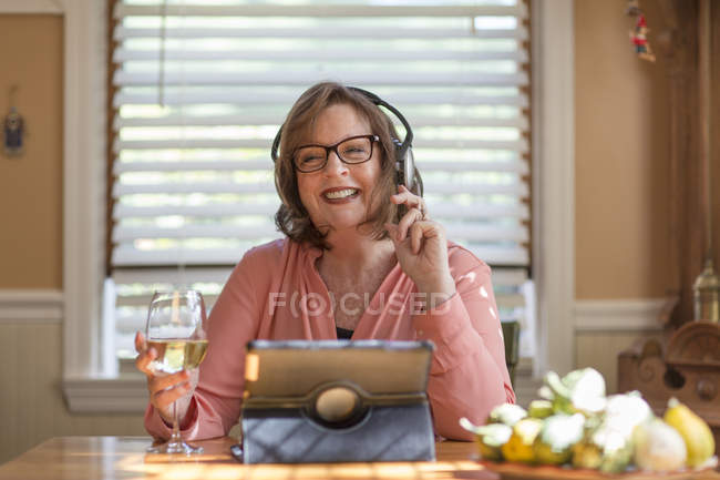 Mulher sênior na mesa da cozinha ouvindo fones de ouvido — Fotografia de Stock