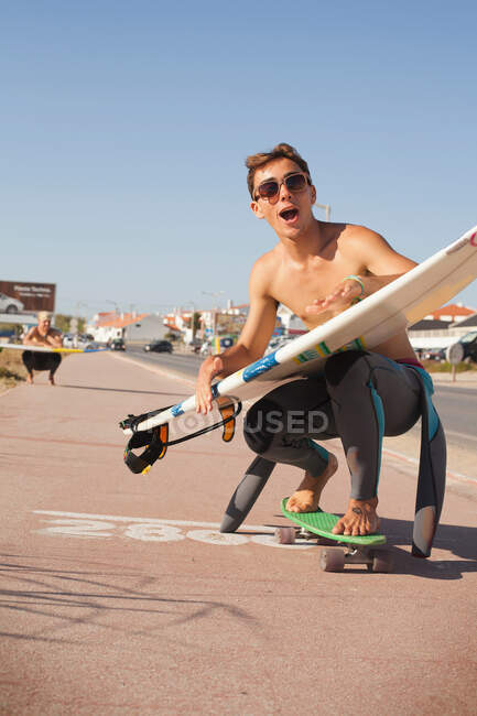 Молодой человек катается на скейтборде вниз по улице, держа доску для серфинга — стоковое фото