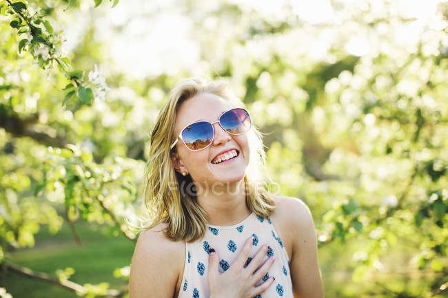 Голова и плечи молодой женщины в солнечных очках, рука о грудь, смотрящей в камеру и смеющейся — стоковое фото