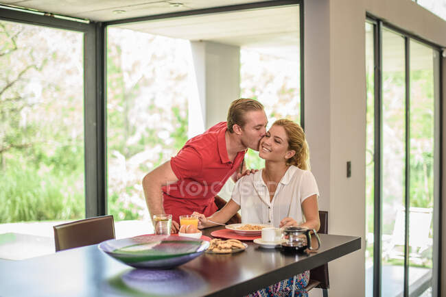 Man kissing girlfriend on cheek at breakfast table — Stock Photo